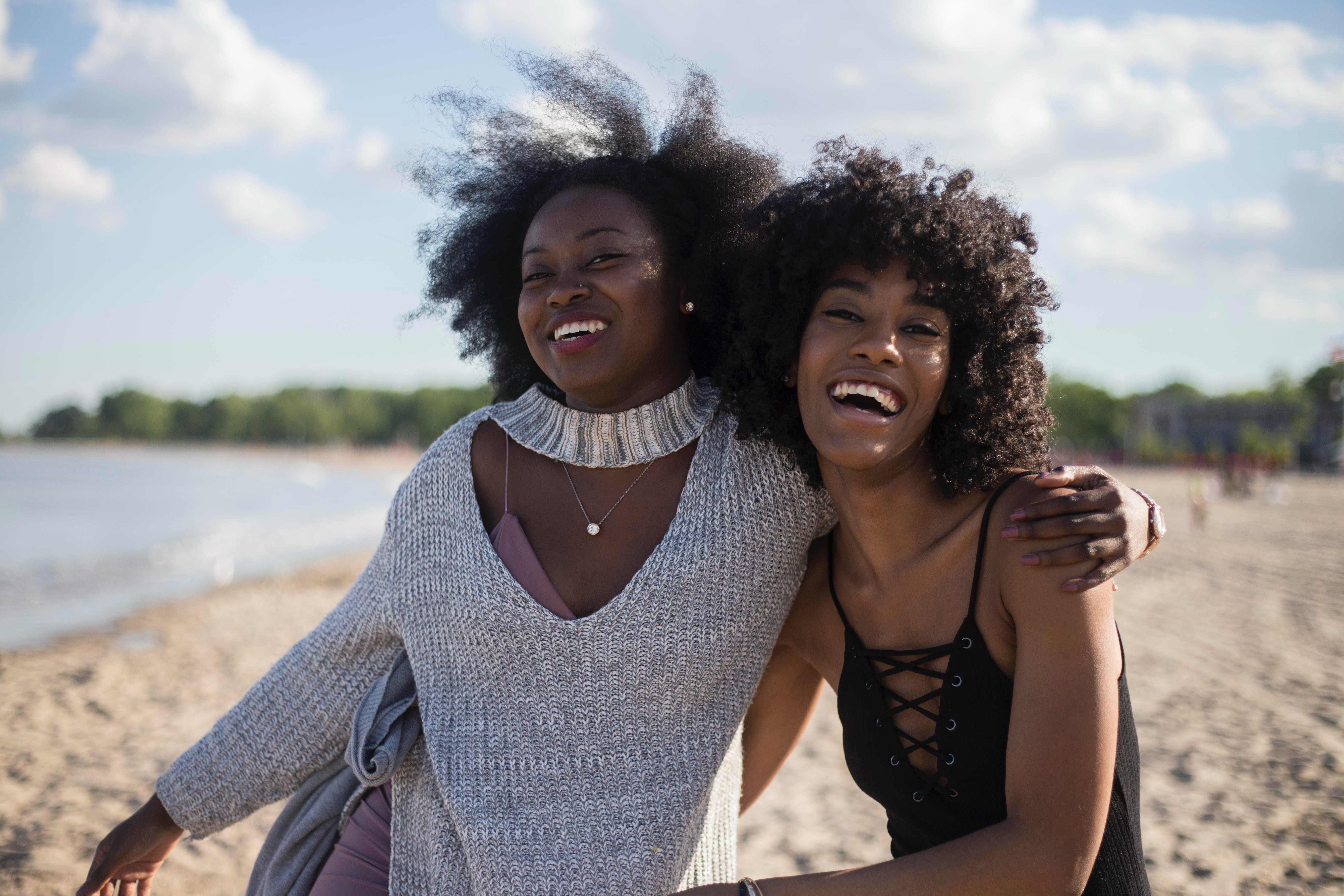 Two young women are arm in arm, smiling on a beach.